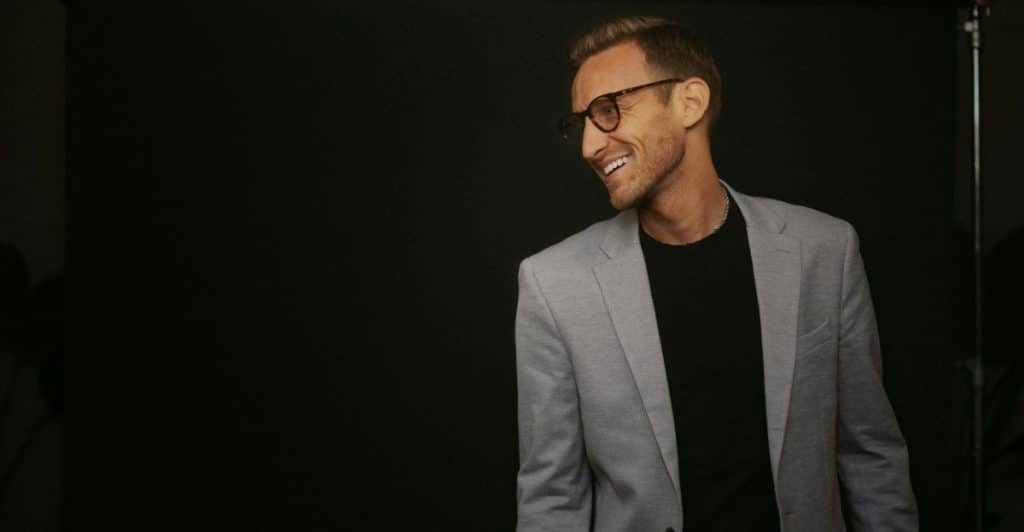 A man in a light grey blazer, black t-shirt, and glasses is smiling and looking to the right against a dark background.