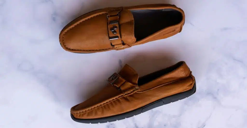 A pair of brown leather loafers with metal buckle accents are placed on a white marble surface.
