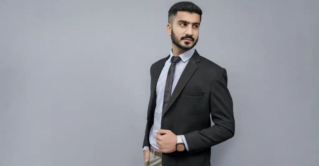 A man in a black suit jacket, light blue shirt, and dark tie with a watch on his wrist is posing against a grey background.
