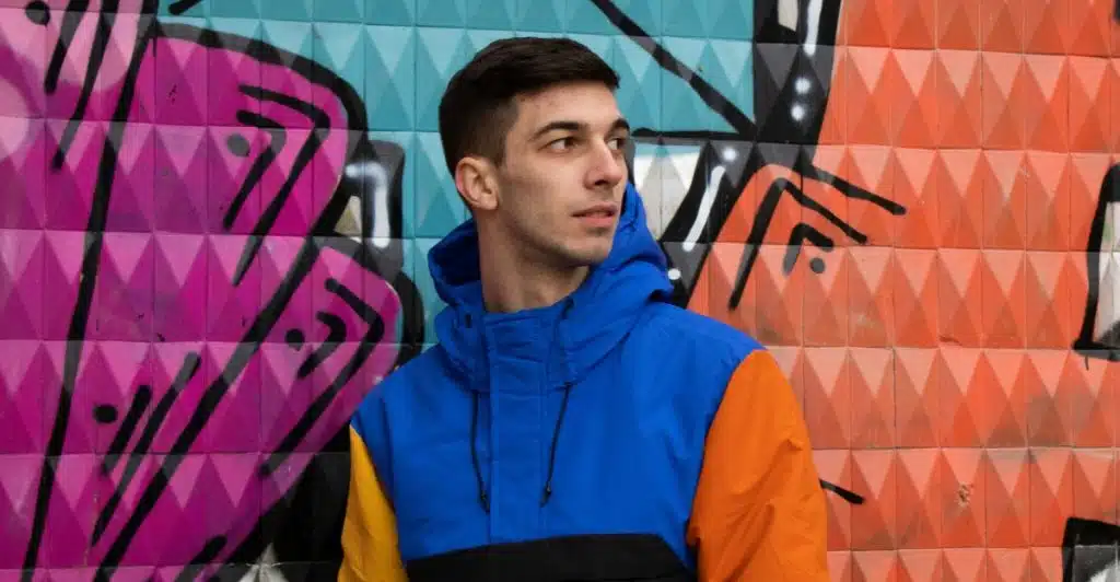 A man in a blue, orange, and black color-block jacket is standing against a colorful graffiti wall.