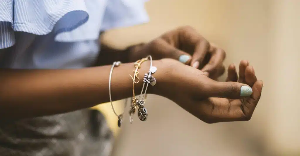 A person with dark skin is adjusting several silver and gold bracelets on their left wrist, with a light blue ruffled sleeve visible.