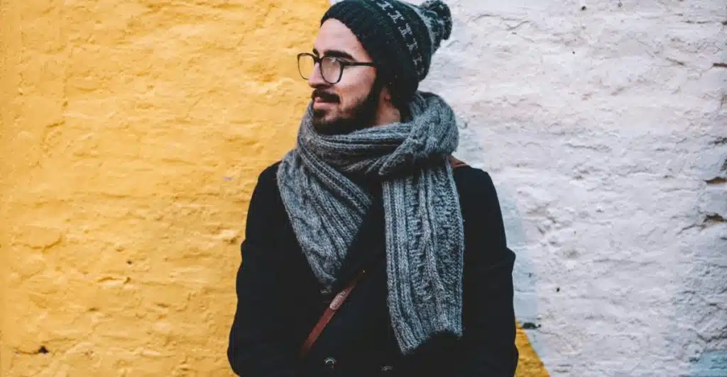 A man wearing a dark beanie, glasses, a grey knitted scarf, and a dark coat is standing outdoors with a yellow and white wall behind him.