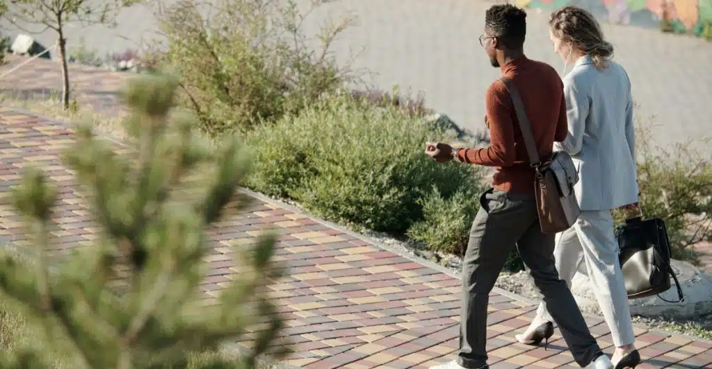 Two people are walking on a paved pathway, one man carrying a brown shoulder bag and the other woman carrying a black tote bag.