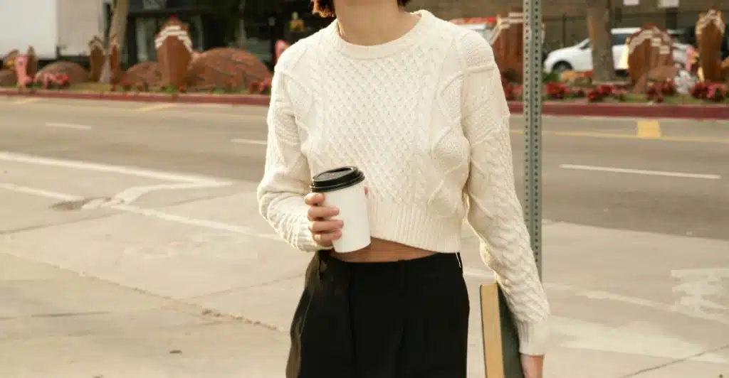 A woman in a white cable-knit sweater and black pants is holding a coffee cup and a book while standing outdoors.