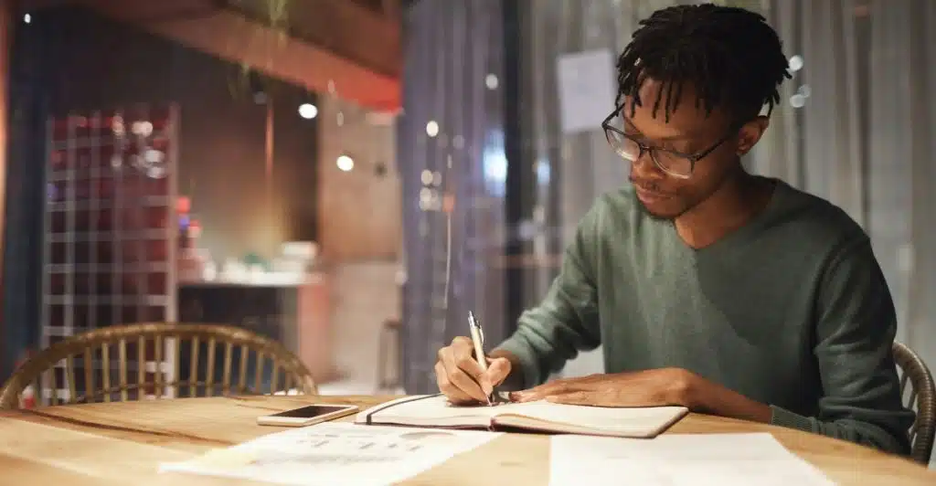 A man with dreadlocks and glasses is sitting at a wooden table, writing in a notebook with a pen.