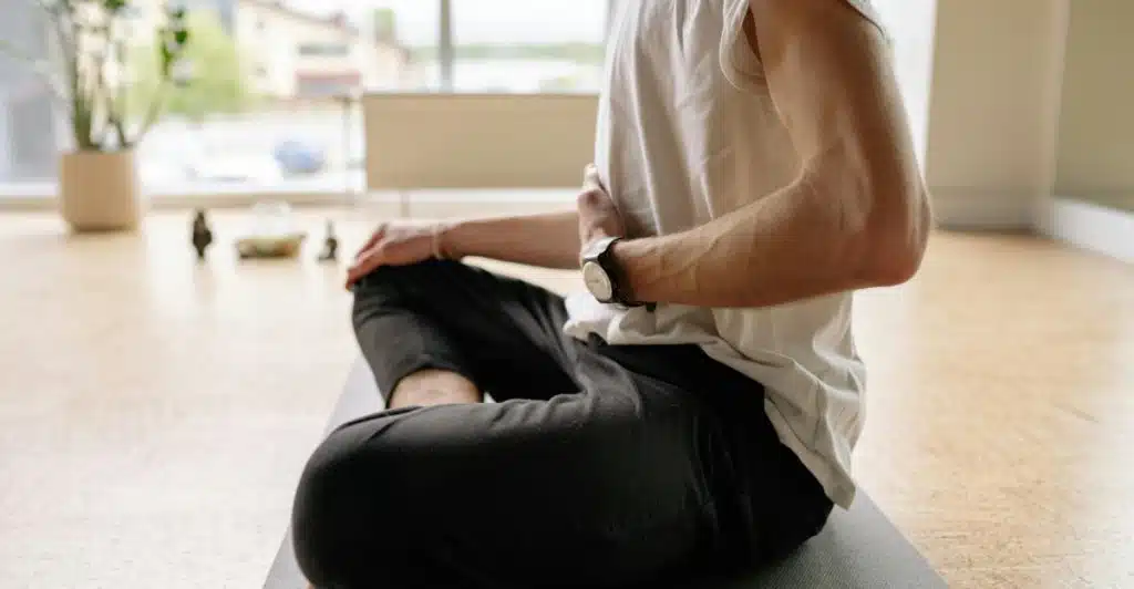 A person sitting cross-legged on a yoga mat in a bright room with one hand on the stomach and the other resting on the knee during a meditation practice