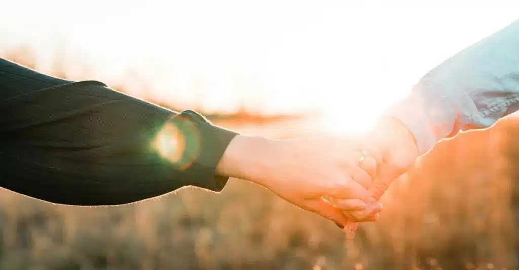 Two people are holding hands in a field with the sun setting in the background.