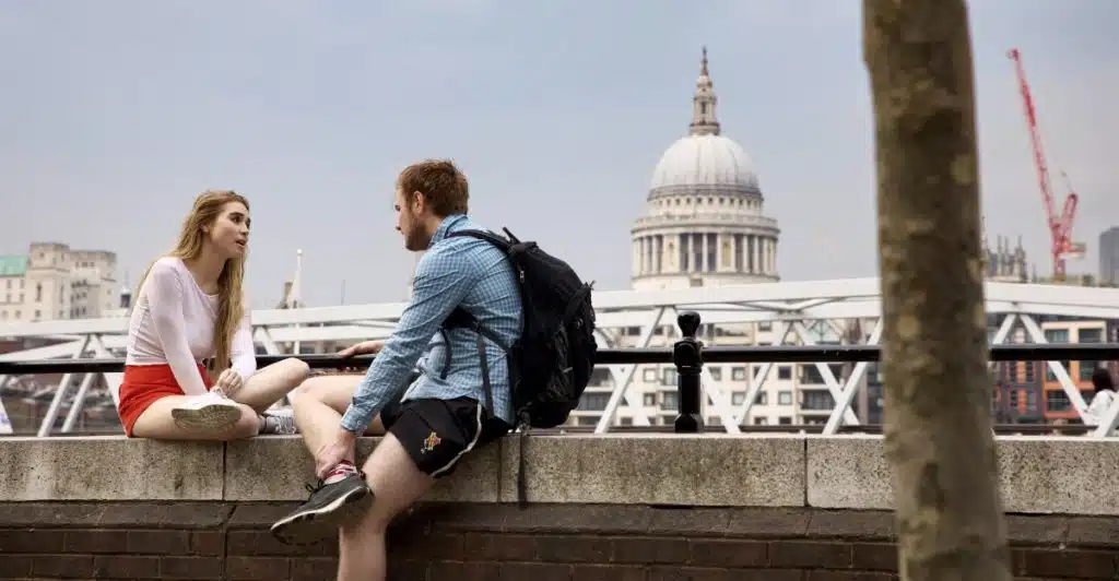 A man with a backpack and a woman are sitting on a stone ledge outdoors, talking to each other with a large building and sky in the background.