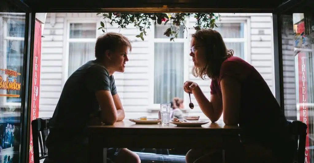 A man and a woman are sitting across from each other at a wooden table in what appears to be a cafe, talking.