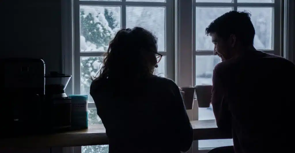 Two people are standing by a window, looking at each other while holding mugs, with a snowy outdoor scene visible through the window.