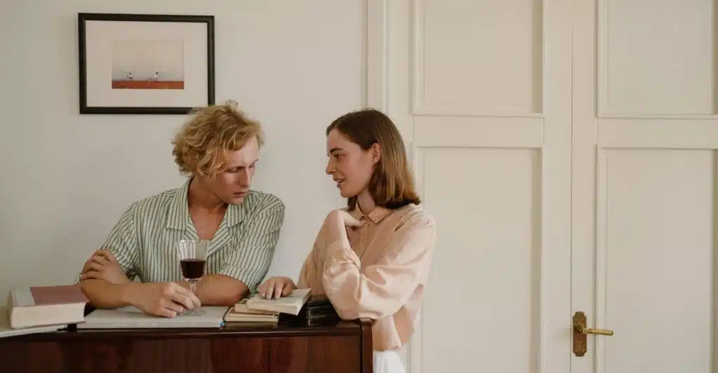 A man is sitting at a piano with a glass of red wine, looking at a woman who is standing next to him and talking.