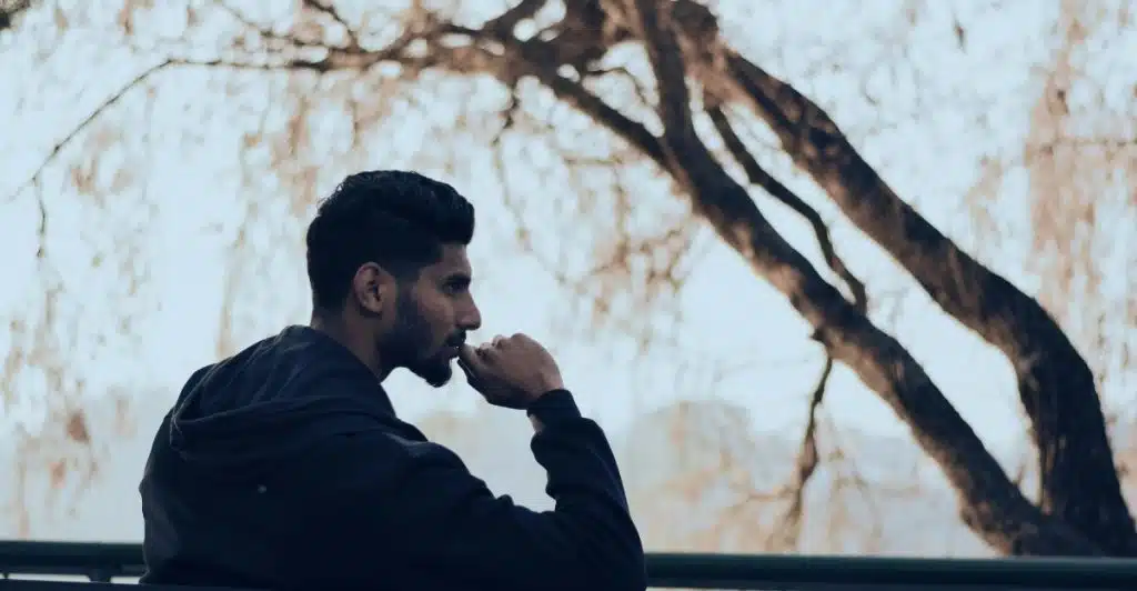 A man sitting alone on a dark bench outdoors, looking to the side with a pensive expression.