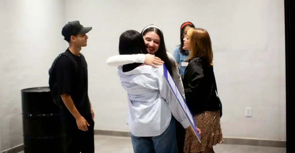 Two women embracing in a hug while other people stand in the background, suggesting a social gathering