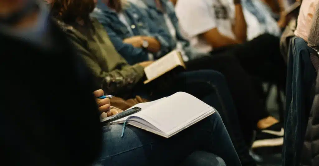 People sitting in rows, some holding books or notebooks, at what appears to be a religious or spiritual gathering