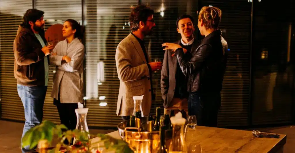A group of people socializing and holding drinks at an evening networking event with bottles and glasses on a table