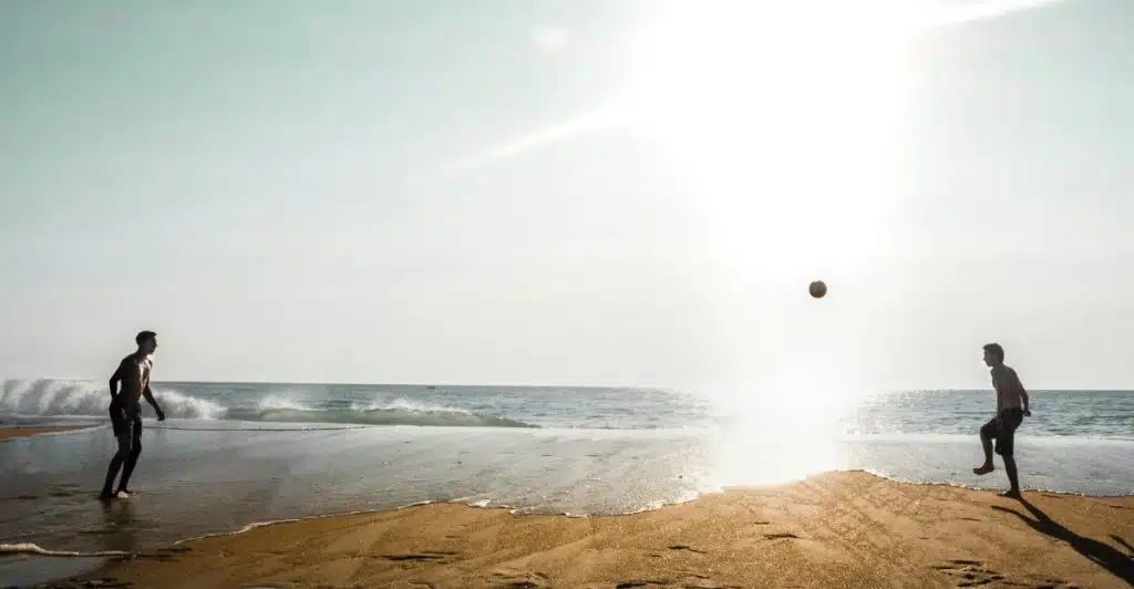 Two men playing soccer on a beach near the ocean with a ball in the air.