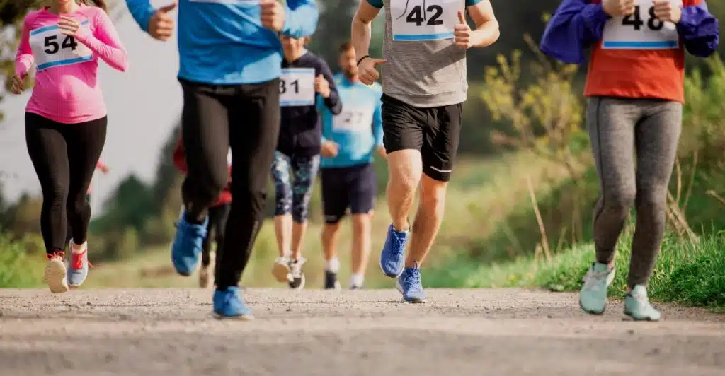 Several people running outdoors on a paved path, participating in a race with numbers pinned to their shirts