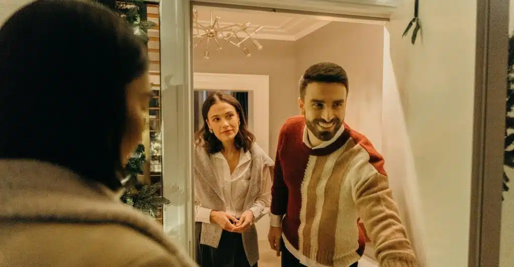 A woman standing at an open doorway, greeting a man and a woman who are standing outside