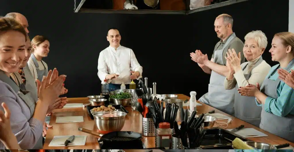 A group of adults participating in a cooking class, with a chef in the center demonstrating, and others standing around a large wooden table with cooking equipment and ingredients