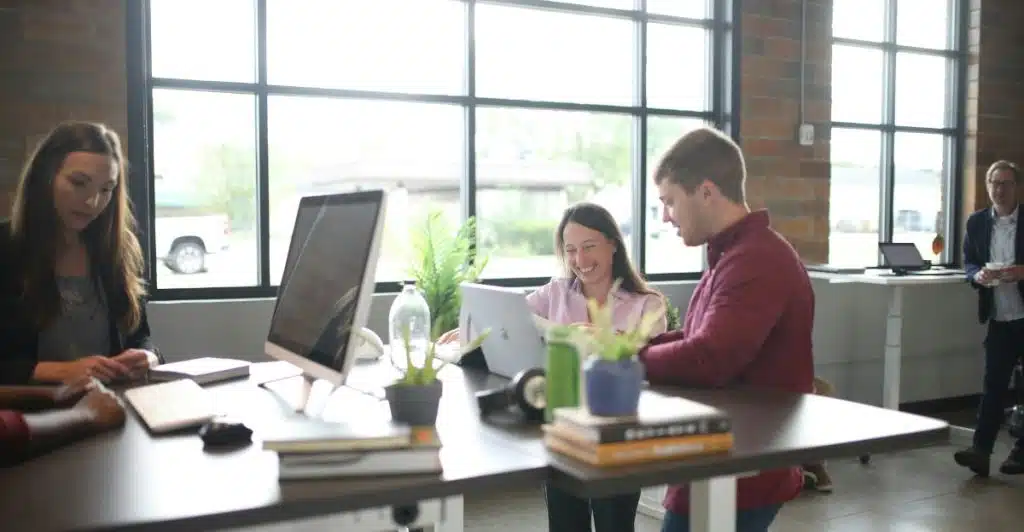 Several people working at standing desks in an open office environment with large windows