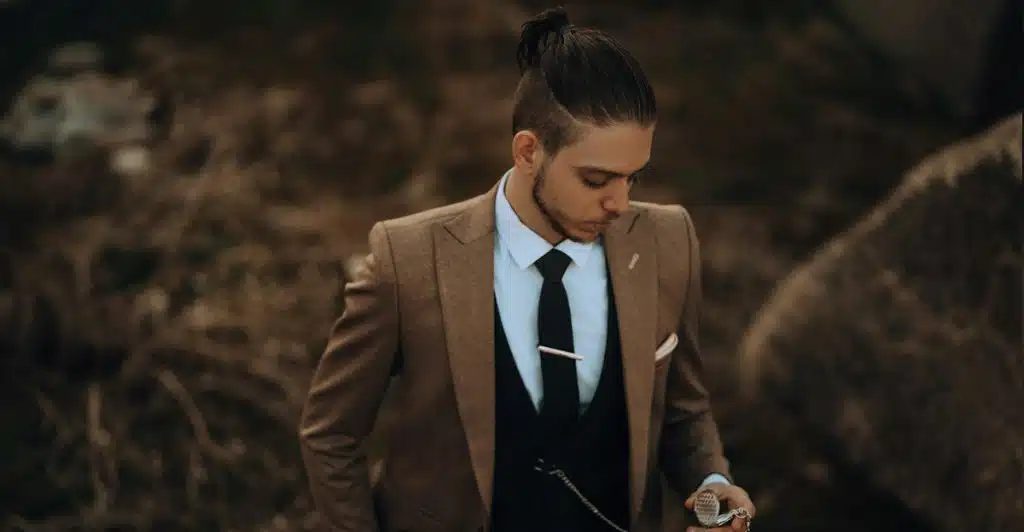 A man in a brown suit and black tie looking down at a pocket watch he is holding.