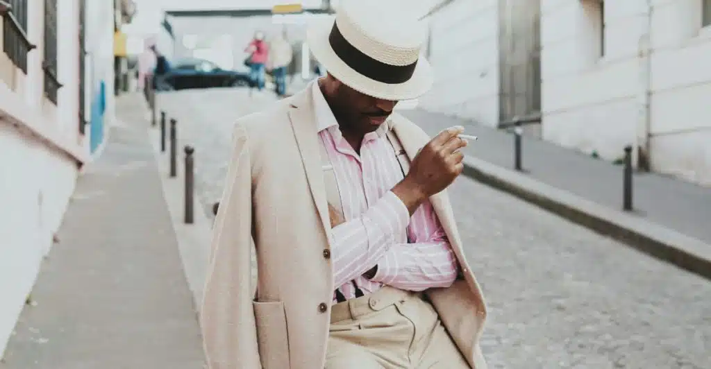 A man in a light-colored suit jacket, pink and white striped shirt, and a straw fedora hat with a black band, holding a cigarette and looking down.