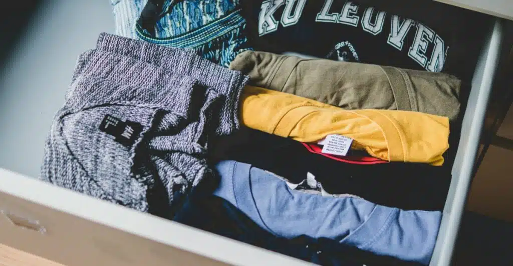 A drawer filled with folded clothes, including a grey sweater, an olive green t-shirt, a yellow t-shirt, and a blue t-shirt.
