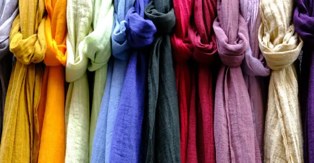 A close-up of a rack displaying numerous folded scarves in various colors, including yellow, orange, light green, blue, and purple.