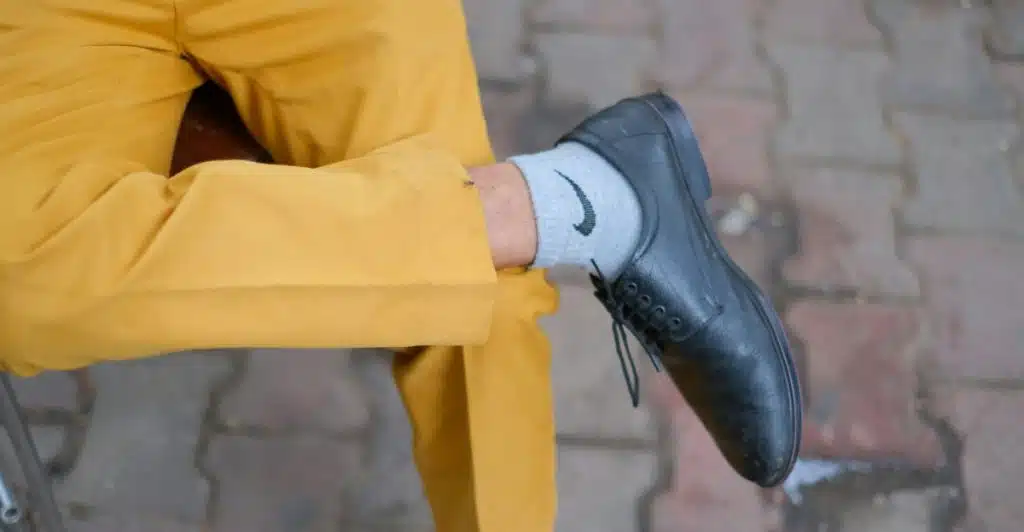 A close-up of a man's legs, one bent, wearing yellow pants, grey socks with a black logo, and black dress shoes, against a brick-patterned ground.