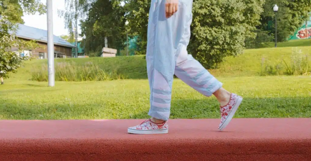 A person walking on a red pathway, wearing light blue and white striped pants and white and red patterned low-top sneakers.