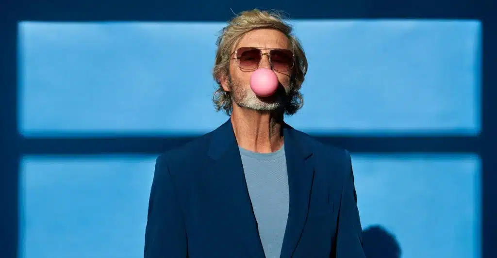 A man with grey hair and sunglasses, wearing a blue suit jacket over a light blue t-shirt, blowing a large pink bubblegum bubble against a blue background with a shadow.