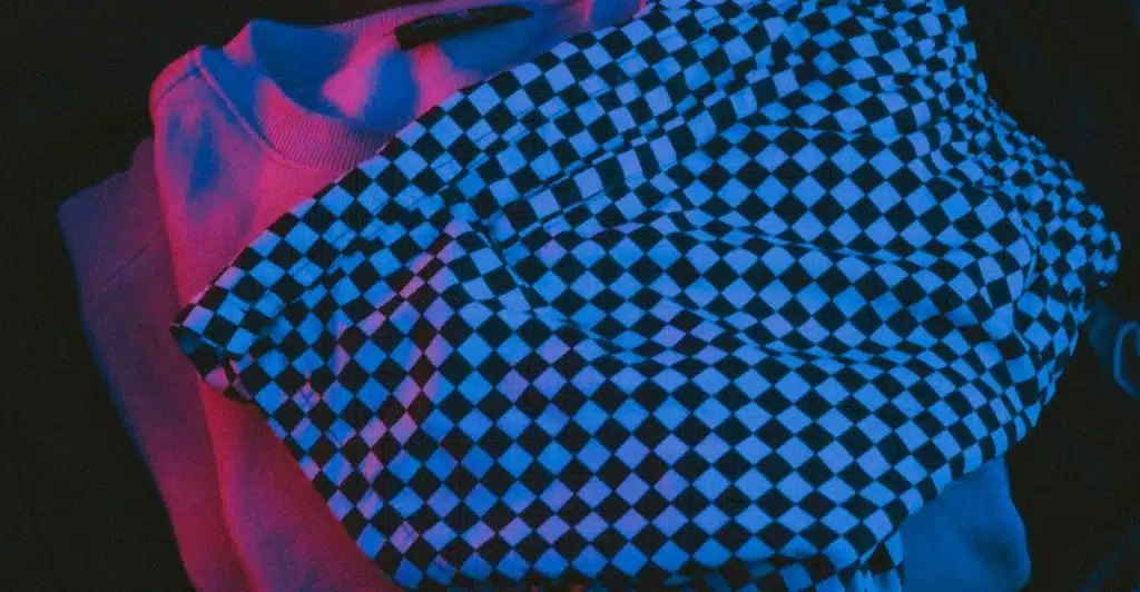A pair of black and white checkered shorts resting on a pinkish-purple t-shirt, under blue and pink lighting.