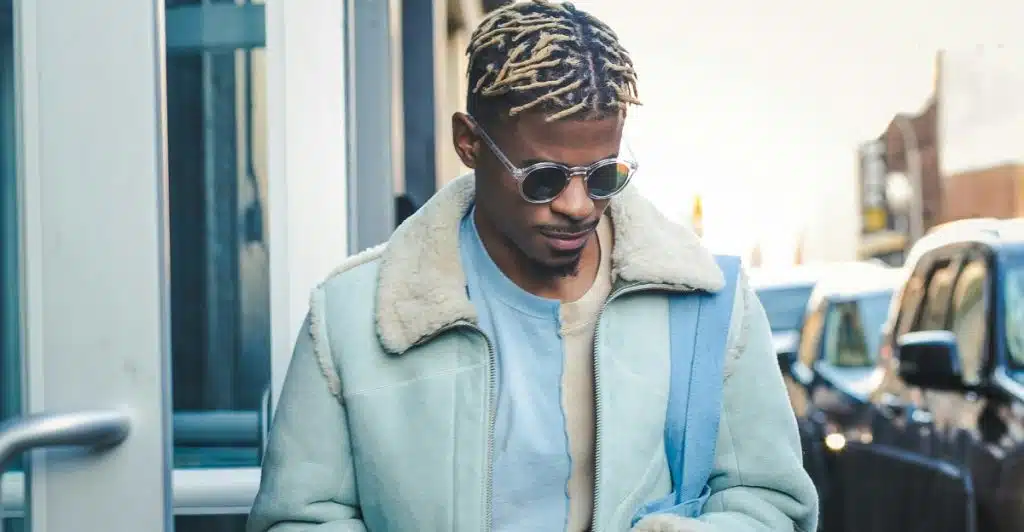 A man with blonde dreadlocks and sunglasses, wearing a light blue and cream shearling-lined jacket over a blue and light blue shirt, looking down outdoors.