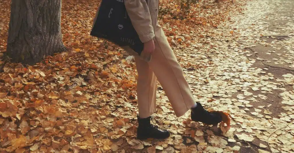 The lower half of a person walking on a path covered in orange and brown autumn leaves, wearing light-colored pants, black boots, and carrying a black tote bag.