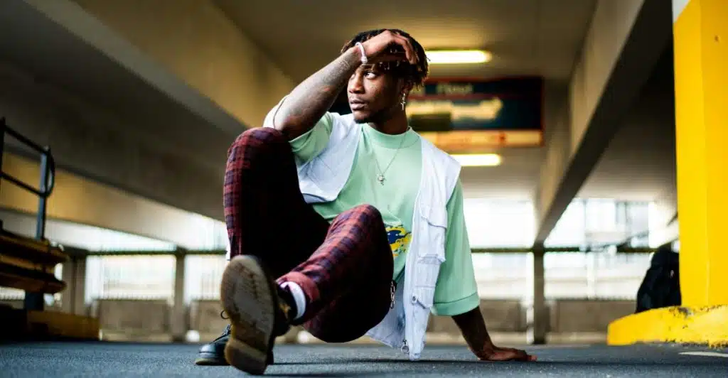 A man with dark skin, wearing a light green t-shirt, a white vest, and red and black plaid pants, sitting with one leg raised in a parking lot.