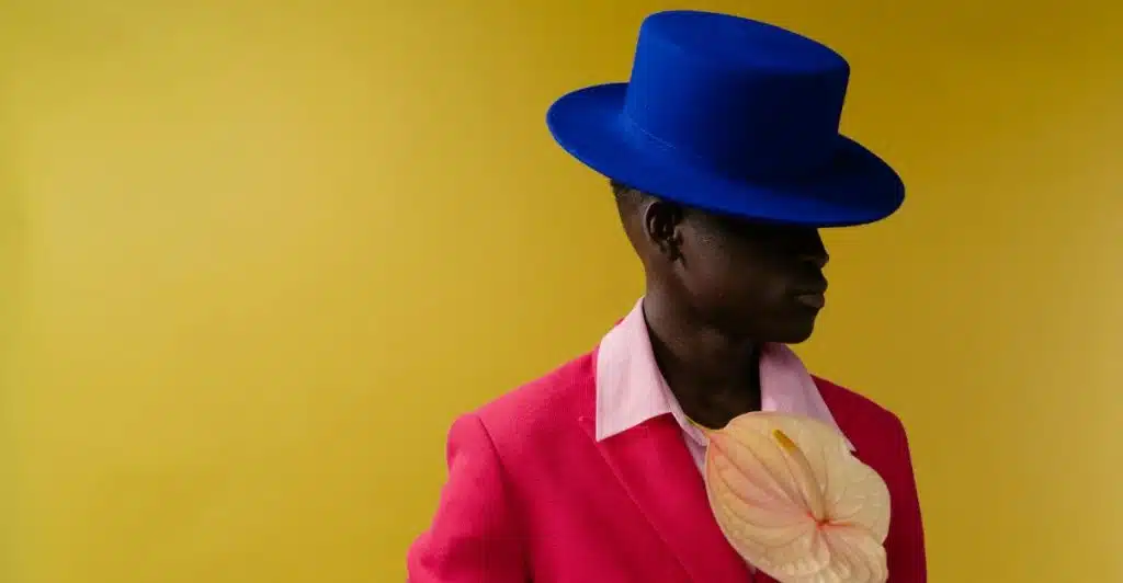 A man with dark skin, wearing a vibrant pink suit jacket over a light pink collared shirt, and a bright blue top hat, holding a light-colored anthurium flower, against a yellow background.