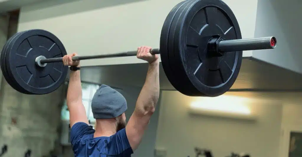 A man from behind, wearing a blue t-shirt and a grey beanie, is lifting a heavy barbell with large black weights overhead in a gym setting.