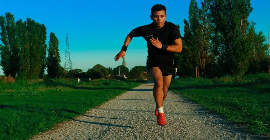 A young man in black shorts and a black t-shirt is sprinting towards the camera on a gravel path, with green grass and tall trees under a bright blue sky.