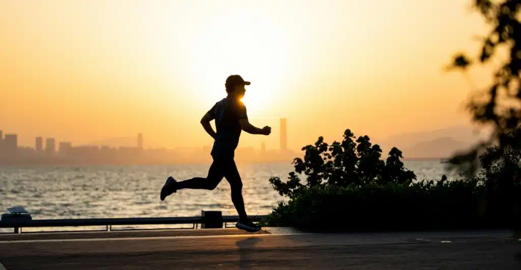 The silhouette of a man wearing a baseball cap is jogging along a paved path next to a body of water during a vibrant sunset, with city buildings visible in the distance.