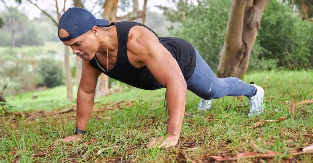 A man in a black tank top, grey pants, and a blue baseball cap is performing a push-up on a grassy area outdoors, with trees and bushes in the background.