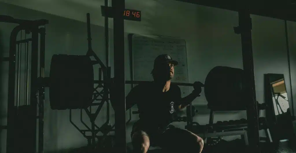 A man in a dark t-shirt and cap is performing a barbell squat, positioned under a squat rack with a clock showing "18:46" in the dimly lit gym.