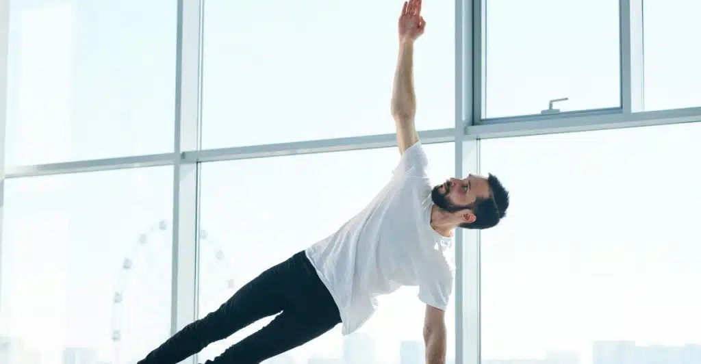 A bearded man in a white t-shirt and black pants is performing a side plank yoga pose on a black mat, with his right arm extended towards the ceiling, against a large window backdrop.