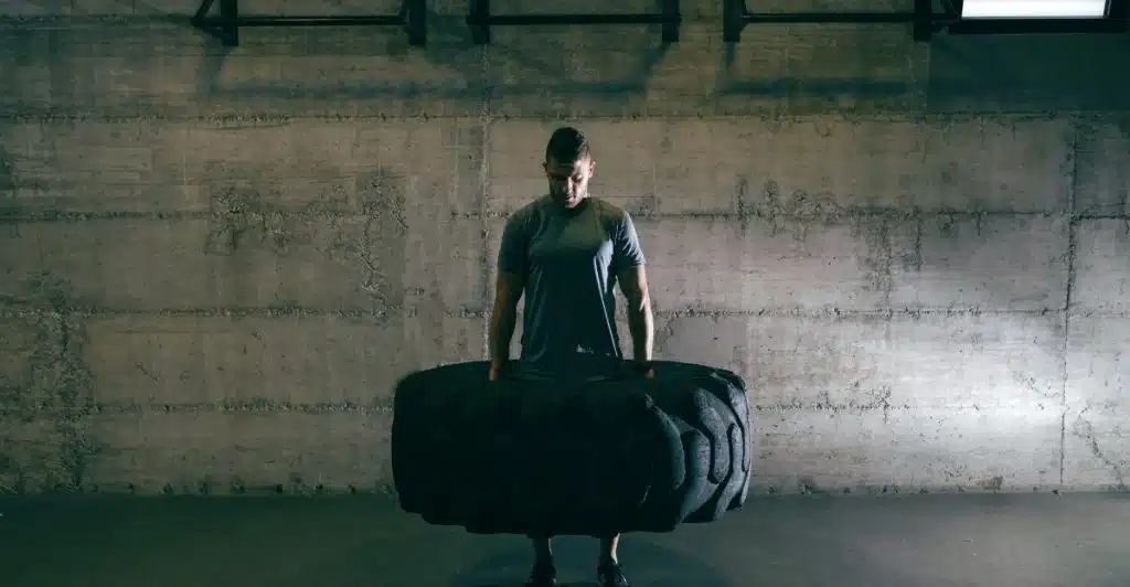 A man in a grey t-shirt is standing in a gym, holding onto the top of a large, heavy-looking black tire that he appears to be lifting or preparing to flip.