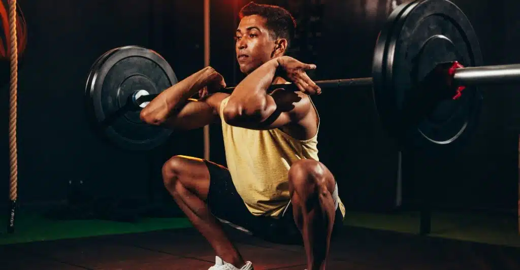 A man in a yellow tank top and black shorts is performing a deep squat with a heavy barbell resting across his upper back in a gym.