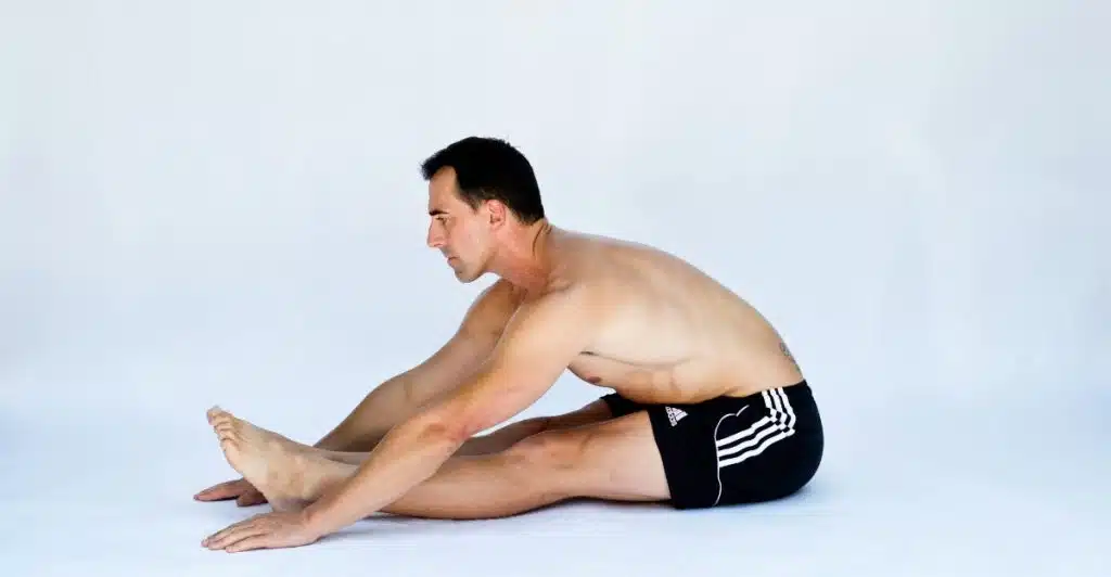 A shirtless man in black shorts is sitting on the floor with his legs extended, reaching forward to touch his toes as part of a stretching exercise.