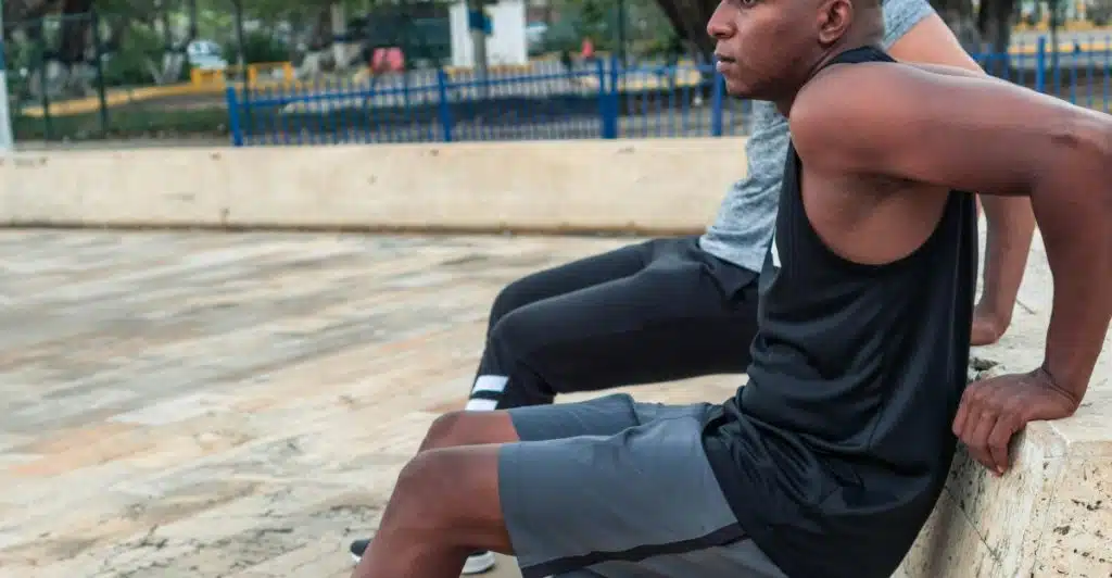 A man in a black tank top and grey shorts is doing triceps dips using a stone ledge in an outdoor park setting.