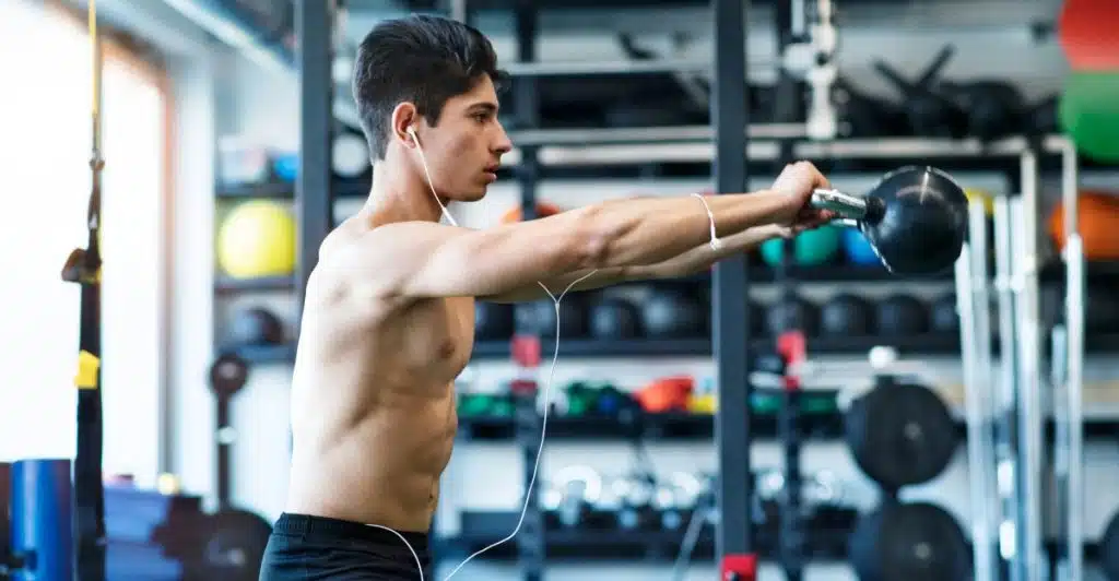 A young man with dark hair, wearing white earphones and black shorts, is performing a kettlebell swing in a gym, holding the kettlebell with both hands extended forward.