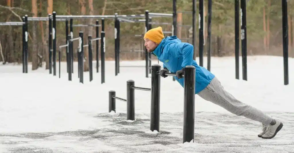 A man wearing a blue jacket, grey pants, and a yellow beanie is doing a push-up exercise on parallel bars in a snowy outdoor workout area surrounded by trees.