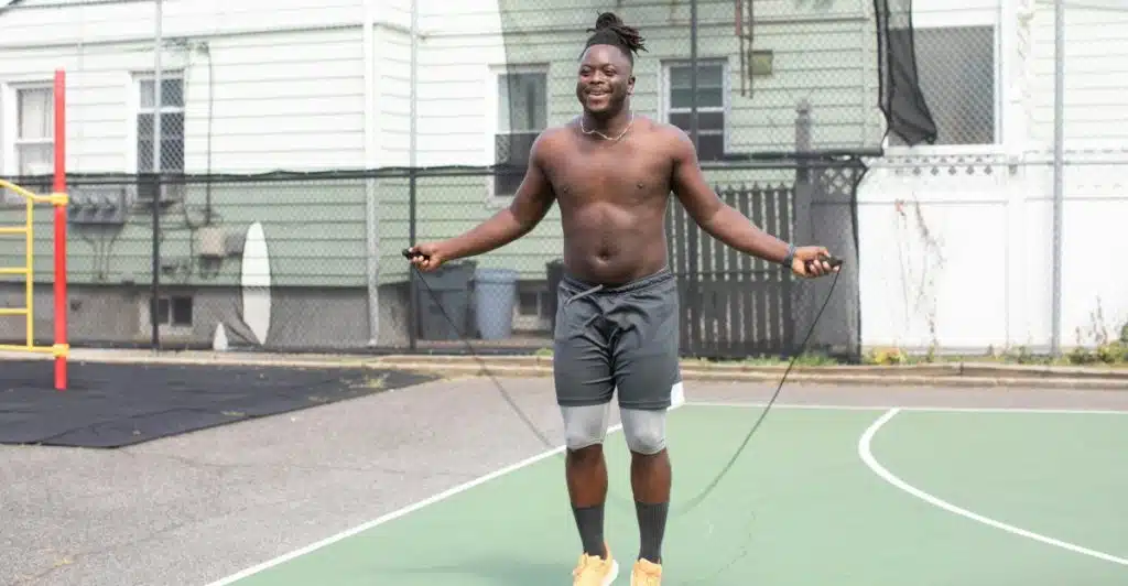A smiling shirtless man with dreadlocks, wearing black shorts and yellow sneakers, is jumping rope on a green outdoor basketball court.