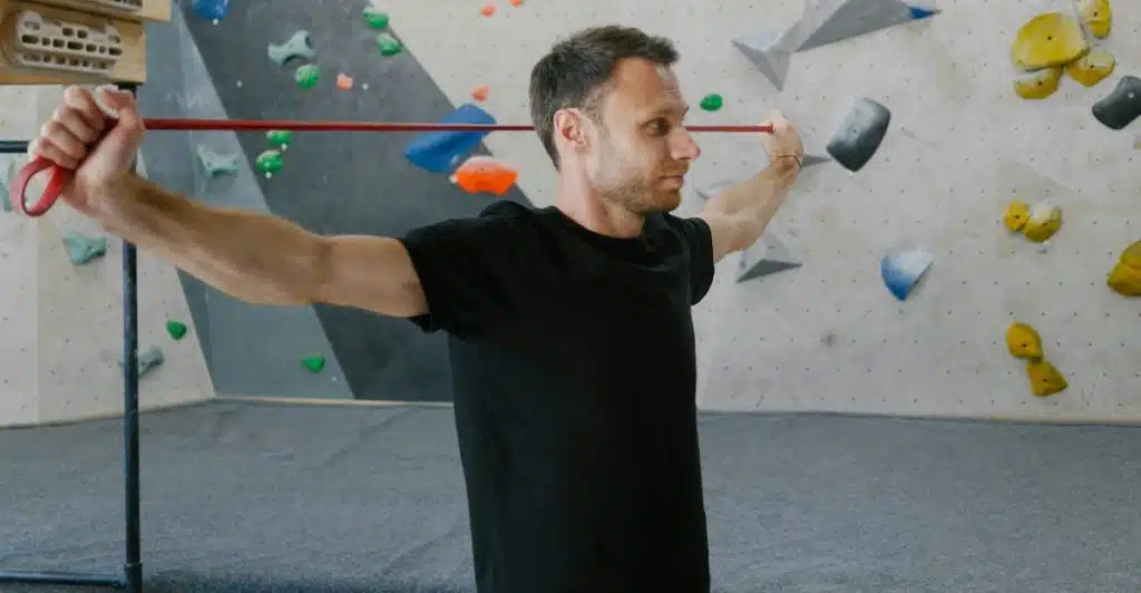 A man in a black t-shirt is standing sideways, stretching a red resistance band across his chest with both hands in a climbing gym with a bouldering wall in the background.